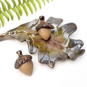 May include: A ceramic oak leaf dish with a glossy green and brown glaze, featuring two matching acorn-shaped ornaments. The acorns have textured, dark brown glazed caps and smooth, matte tan bases, mirroring the natural form of forest foliage.