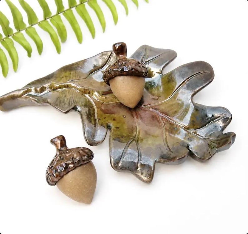 May include: A ceramic oak leaf dish with a glossy green and brown glaze, featuring two matching acorn-shaped ornaments. The acorns have textured, dark brown glazed caps and smooth, matte tan bases, mirroring the natural form of forest foliage.