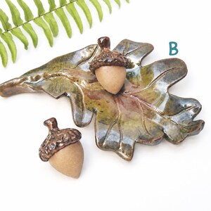 May include: A ceramic oak leaf-shaped dish with a glossy green and brown glaze, accompanied by two acorn-shaped ceramic pieces with textured brown caps and matte tan bases. The letter B is printed in blue near the top right of the leaf dish.