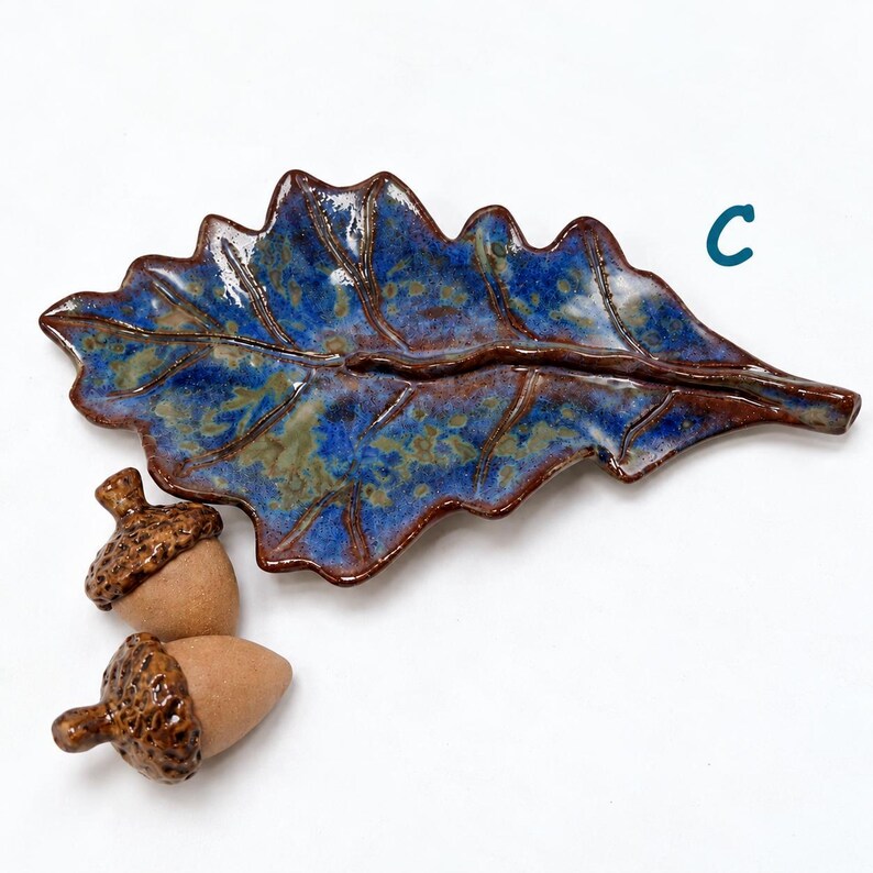 May include: A ceramic leaf-shaped dish with a glossy blue and brown glaze, featuring a central vein and scalloped edges, accompanied by two small ceramic acorns with textured brown caps and smooth tan bodies. The letter C is visible in blue.
