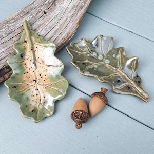 May include: Two ceramic dishes shaped like oak leaves with a glossy green and beige glaze featuring dark speckles, accompanied by two small brown ceramic acorns with textured caps, arranged together on a light blue wooden surface.