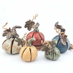 May include: A collection of five ceramic pumpkin sculptures in various colors including light blue, orange, tan, and dark green. Each pumpkin features a textured surface, a sculpted ceramic leaf, and a decorative copper wire stem.