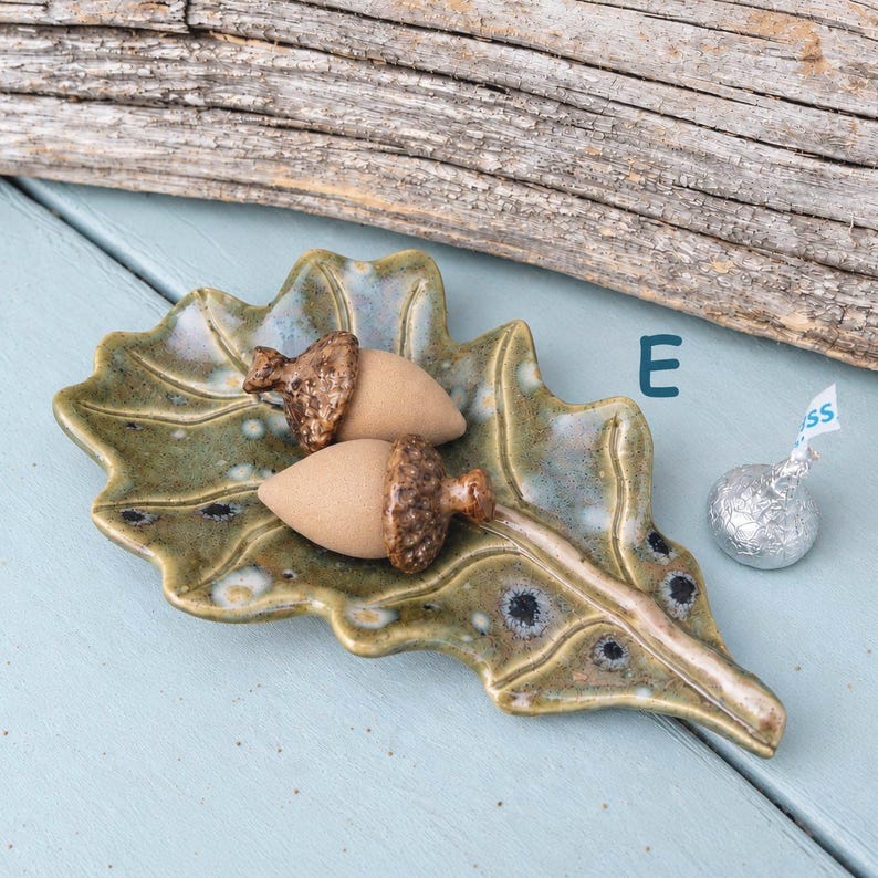 May include: A ceramic leaf-shaped dish in mottled green with two tan acorn-shaped objects featuring textured brown caps resting on top. The letter E is printed in blue near the dish, and a silver foil-wrapped chocolate candy sits nearby.