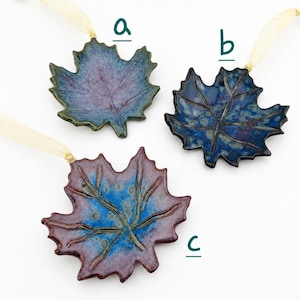 Op de afbeelding: Drie keramische esdoornbladornamenten met cr&egrave;mekleurige linten. Ornament a is paars en blauw, ornament b is donkerblauw met spikkels en ornament c is een mix van blauw en roodachtig paars. Elk blad heeft reli&euml;faders.