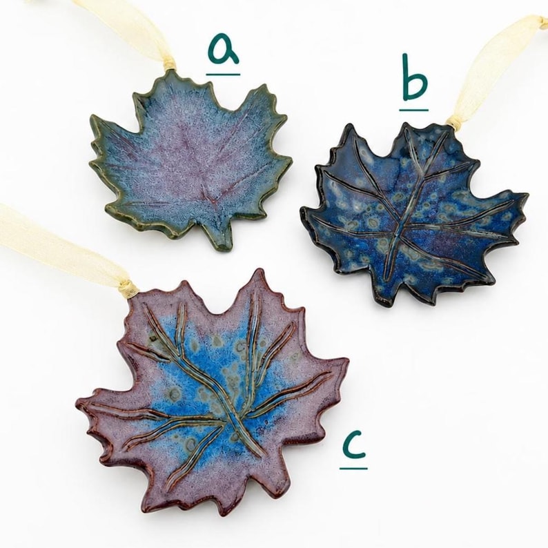 Op de afbeelding: Drie keramische esdoornbladornamenten met cr&egrave;mekleurige linten. Ornament a is paars en blauw, ornament b is donkerblauw met spikkels en ornament c is een mix van blauw en roodachtig paars. Elk blad heeft reli&euml;faders.