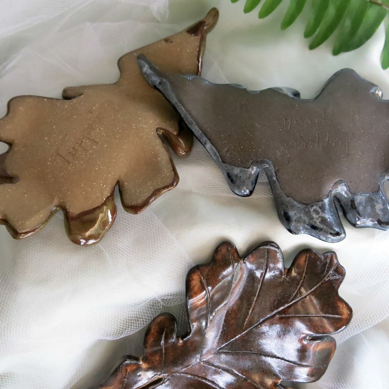 May include: Three ceramic dishes shaped like oak leaves with glossy finishes. One dish is a light tan, one is a dark charcoal gray, and one is a deep brown with visible vein detailing. Each piece features a smooth, glazed surface texture.