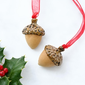 May include: Two ceramic acorn ornaments featuring smooth, matte tan nut bodies and glossy, textured brown caps. Each acorn is attached to a thin, translucent red ribbon for hanging, designed with a realistic, detailed finish for seasonal decor.