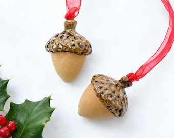 May include: Two ceramic acorn ornaments featuring smooth, matte tan nut bodies and glossy, textured brown caps. Each acorn is attached to a thin, translucent red ribbon for hanging, designed with a realistic, detailed finish for seasonal decor.