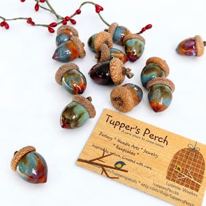May include: A collection of ceramic acorns with glossy, multi-coloured glazes in shades of blue, brown, and red, each topped with a textured brown cap. A rectangular wooden business card for Tupper's Perch with text and a birdcage graphic.