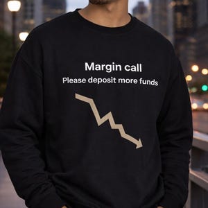 May include: Black sweatshirt with the text "Margin call Please deposit more funds" and a downward trending arrow graphic. The person is wearing blue jeans and standing on a bridge.