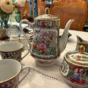 May include: A porcelain tea set with a colorful floral and figural design. The set includes a teapot, sugar bowl, creamer, and two teacups. The teapot has a gold-colored lid and handle. The teacups have gold rims. The set is displayed on a white tablecloth.