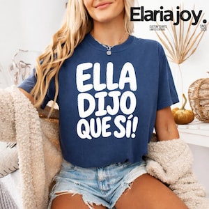 May include: A navy blue short-sleeved t-shirt featuring the white text ELLA DIJO QUE SÍ! printed in a bold, rounded font on the chest. The shirt has a crew neckline and is paired with light blue denim shorts and a fuzzy cream-colored cardigan.
