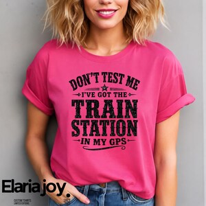 Peut inclure: Un t-shirt magenta avec le texte "DON'T TEST ME I'VE GOT THE TRAIN STATION IN MY GPS" en noir. Le t-shirt a des manches retroussées et est porté par une personne.