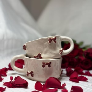 Peut inclure: Deux tasses en céramique blanc cassé empilées, avec des nœuds rouges. Les tasses ont une surface texturée et des anses incurvées. Des pétales de roses rouges sont dispersés autour des tasses, avec un arrière-plan flou de roses et de tissu blanc.