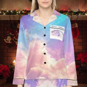 May include: A pajama set featuring a pastel sky and cloud design. The long-sleeved top has black buttons and trim, with a pocket that reads "Your Name" and "Skydaddy's girl". The pajamas are made of a soft, silky material.