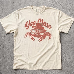 Könnte beinhalten: Cremefarbenes T-Shirt mit roter Grafik einer Krabbe mit Cowboyhut und dem Schriftzug "Yee Claw" in Rot. Das Shirt hat einen klassischen Rundhalsausschnitt und kurze Ärmel.