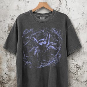 Könnte beinhalten: Ein anthrazitfarbenes Kurzarm-T-Shirt mit einem hellvioletten Grafikdruck einer Spinne in einem Spinnennetz. Das T-Shirt hat einen Rundhalsausschnitt, hängt an einem Holzkleiderbügel und weist eine verwaschene, graue Stoffstruktur auf.