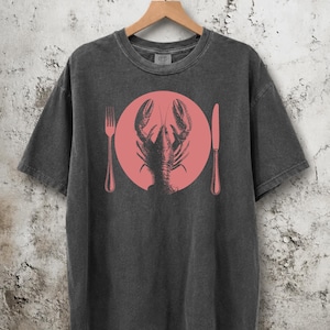 Könnte beinhalten: Ein anthrazitfarbenes Kurzarm-T-Shirt mit einem Grafikdruck eines schwarzen Hummers, der in einem soliden rosa Kreis zentriert ist, flankiert von einer schwarzen Gabel links und einem schwarzen Messer rechts auf dem Rundhals-Kleidungsstück.