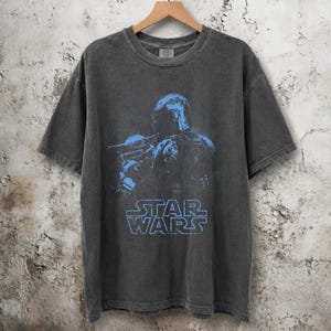 Puede incluir: Una camiseta de manga corta gris carb&oacute;n con un estampado gr&aacute;fico azul de un personaje con casco que sostiene a una peque&ntilde;a criatura. El texto STAR WARS est&aacute; impreso en azul debajo del gr&aacute;fico. La camiseta tiene cuello redondo y acabado desgastado.