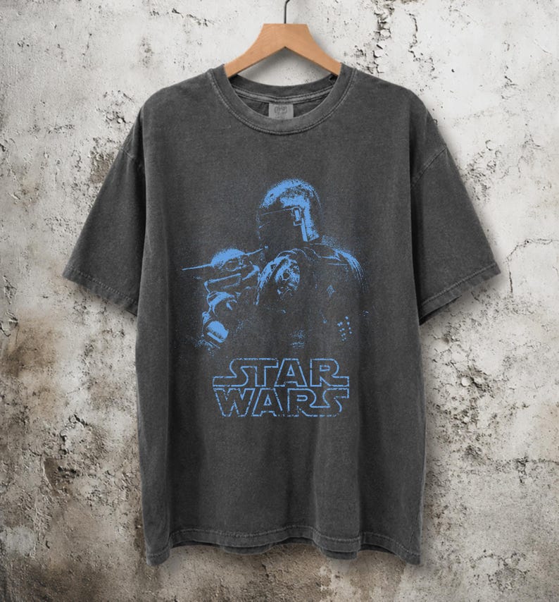 Puede incluir: Una camiseta de manga corta gris carb&oacute;n con un estampado gr&aacute;fico azul de un personaje con casco que sostiene a una peque&ntilde;a criatura. El texto STAR WARS est&aacute; impreso en azul debajo del gr&aacute;fico. La camiseta tiene cuello redondo y acabado desgastado.