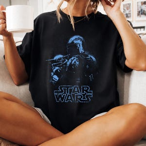 Puede incluir: Una camiseta negra de manga corta con un estampado gr&aacute;fico azul de un personaje con casco que sostiene a una peque&ntilde;a criatura. Debajo de la ilustraci&oacute;n, el texto STAR WARS est&aacute; impreso en azul con una fuente de estilo bloque desgastado.