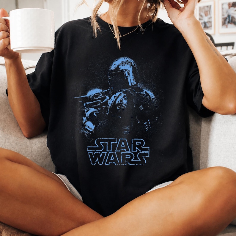 Puede incluir: Una camiseta negra de manga corta con un estampado gr&aacute;fico azul de un personaje con casco que sostiene a una peque&ntilde;a criatura. Debajo de la ilustraci&oacute;n, el texto STAR WARS est&aacute; impreso en azul con una fuente de estilo bloque desgastado.