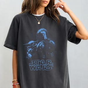 Puede incluir: Una camiseta de manga corta en color gris carb&oacute;n con un estampado gr&aacute;fico azul desgastado de un personaje con casco y una peque&ntilde;a criatura. El texto STAR WARS aparece impreso en azul debajo de las figuras. Tiene cuello redondo.