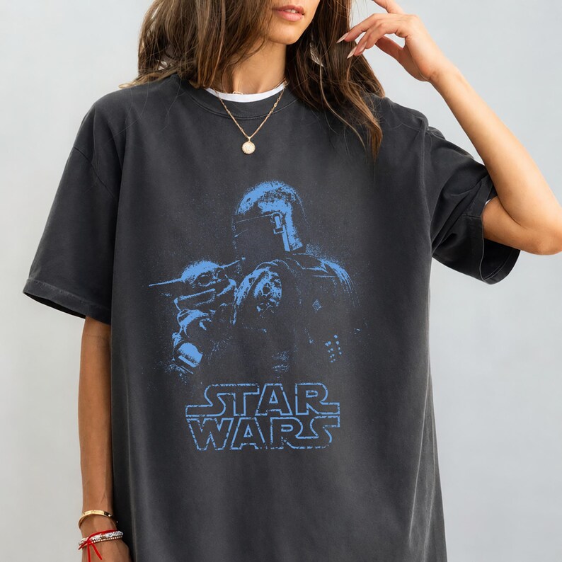 Puede incluir: Una camiseta de manga corta en color gris carb&oacute;n con un estampado gr&aacute;fico azul desgastado de un personaje con casco y una peque&ntilde;a criatura. El texto STAR WARS aparece impreso en azul debajo de las figuras. Tiene cuello redondo.