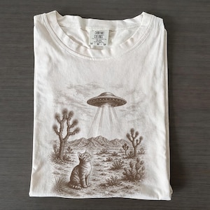 Könnte beinhalten: Cremefarbenes T-Shirt mit einem sepiafarbenen Aufdruck einer Katze, die zu einer UFO über einer Wüstenlandschaft mit Kakteen und Bergen aufblickt. Das Shirt hat ein Comfort Colors-Etikett.