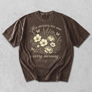 Könnte beinhalten: Braunes T-Shirt mit floralem Design und dem Text "His mercies are new every morning" und "Lamentations 3:22-23". Das Design zeigt Blumen, Schmetterlinge und Blätterwerk im Vintage-Stil.