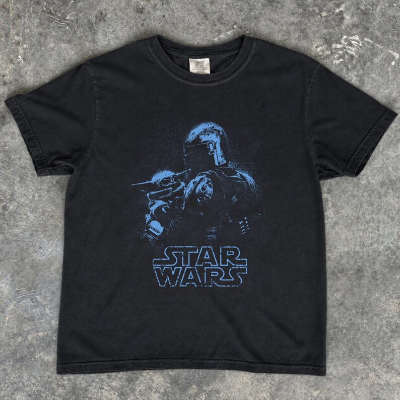 Puede incluir: Una camiseta negra de manga corta con un estampado gr&aacute;fico azul de un personaje con casco que sostiene a una peque&ntilde;a criatura. Debajo de la ilustraci&oacute;n, el texto STAR WARS est&aacute; impreso en un azul a juego con una fuente de bloque desgastada.