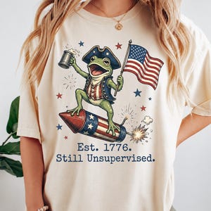 Könnte beinhalten: Ein cremefarbenes T-Shirt mit einer Grafik eines grünen Frosches, der einen Dreispitz und eine Weste trägt, einen Bierkrug und eine amerikanische Flagge hält und auf einer Feuerwerksrakete reitet. Der Text lautet Est. 1776. Still Unsupervised.