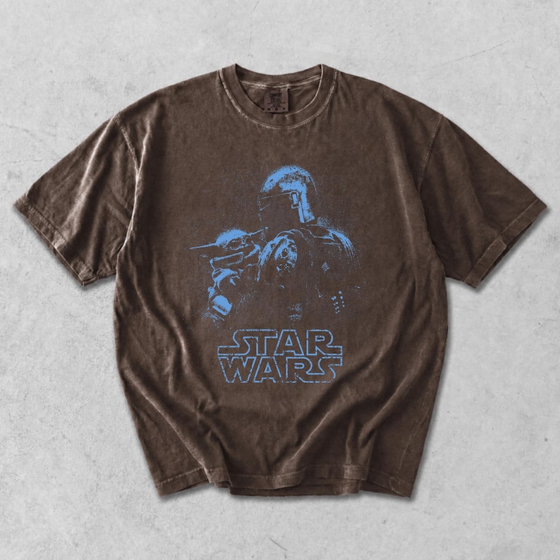 Puede incluir: Una camiseta marr&oacute;n de manga corta con una textura desgastada y deste&ntilde;ida. El centro presenta un estampado gr&aacute;fico en azul claro de un personaje con casco que sostiene a una peque&ntilde;a criatura, con el texto STAR WARS en azul claro debajo.
