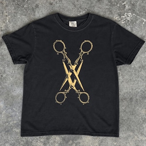 Könnte beinhalten: Ein schwarzes Kurzarm-T-Shirt mit einem mittigen Grafikdruck von zwei gekreuzten, vintage-artigen Scheren in Hellgelb. Die Scheren haben verzierte Griffe und spitze Klingen, die ein symmetrisches, X-förmiges Design auf dem Kleidungsstück bilden.