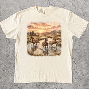 Könnte beinhalten: Ein cremefarbenes Kurzarm-T-Shirt mit einem Grafikdruck von drei braunen Pferden, die durch einen flachen Bach laufen. Der Hintergrund zeigt eine Landschaft mit grasbewachsenen Feldern, fernen Bergen und einem Sonnenuntergangshimmel mit orangefarbenen Wolken.