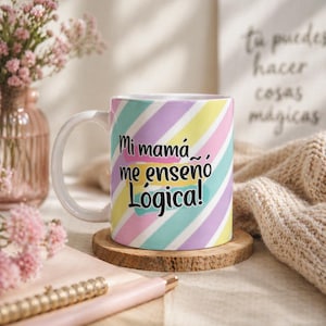 Puede incluir: Una taza de cerámica blanca con un diseño de rayas pastel en tonos rosa, amarillo y azul. La taza presenta el texto en español "Mi mamá me enseñó Lógica!". La taza está sobre un posavasos de madera.