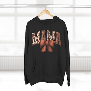 Puede incluir: Sudadera con capucha negra con la palabra "MAMA" en una mezcla de estampado animal y letras con purpurina. Un gran lazo marrón está centrado debajo de la palabra. La sudadera tiene una capucha con cordón y un bolsillo delantero.