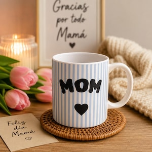Puede incluir: Una taza de cerámica blanca con un patrón de rayas verticales azul claro, que presenta la palabra MOM en letras negras en negrita sobre un corazón negro sólido. La taza tiene un asa curva y se asienta sobre un posavasos redondo trenzado.