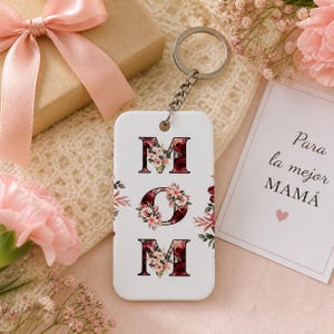 May include: A white rectangular keychain with the word "MOM" in burgundy letters decorated with floral designs. The keychain is attached to a silver keyring. A gift box and a card with text are in the background.