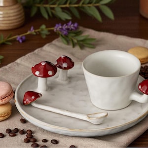 Op de afbeelding: Een witte keramische kop en schotel set met paddenstoel accenten. De set bevat een kopje, een schotel met drie paddenstoel beeldjes en een lepel. Twee macarons en koffiebonen zijn ook aanwezig.