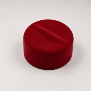 Könnte beinhalten: Ein einfarbiger roter Fez-Hut mit zylindrischer Form und flacher Oberseite. Der Hut hat eine dezente Einkerbung über die Oberseite. Der Hut besteht aus einem filzartigen Material und steht vor einem weißen Hintergrund.