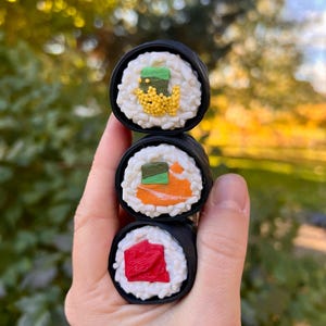 Puede incluir: Tres rollos de sushi con arroz blanco, rellenos coloridos y envolturas de algas marinas negras están apilados verticalmente. El rollo superior tiene aderezos verdes y amarillos, el rollo del medio tiene rellenos naranjas y verdes, y el rollo inferior tiene relleno rojo.