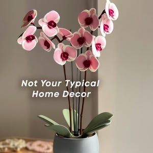 May include: A faux orchid in a grey ceramic pot, featuring pink and white flowers and green leaves. The image includes the text "Not Your Typical Home Decor". A decorative piece for the home.