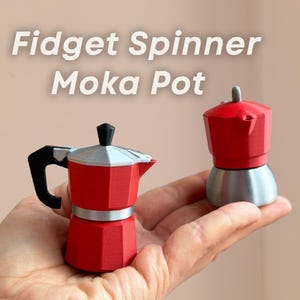 Könnte beinhalten: Zwei rote und silberne Fidget Spinner Moka-Kannen. Eine wird in einer Hand gehalten, die andere daneben. Der Text "Fidget Spinner Moka Pot" steht darüber. Die Moka-Kannen haben einen Griff und ein silbernes Band.