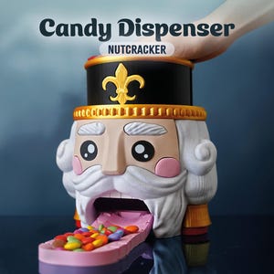 Könnte beinhalten: Ein neuartiger Süßigkeiten-Spender in Form eines Nussknacker-Soldaten. Der Spender ist schwarz, gold und beige mit weißem Bart und rosa Wangen. Der Text "Candy Dispenser NUTCRACKER" steht darüber. Bunte Süßigkeiten werden aus dem Mund abgegeben.