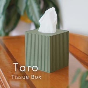 Op de afbeelding: Een vierkante, saliegroene tissuebox met verticale ribbels, met een stapel witte tissues. De tekst "Taro Tissue Box" staat op het houten oppervlak voor de doos.