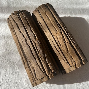 May include: Two cylindrical wooden containers with a textured, bark-like appearance. The containers are a medium brown color and appear to be made of carved wood. The containers are lying on a white, textured surface.