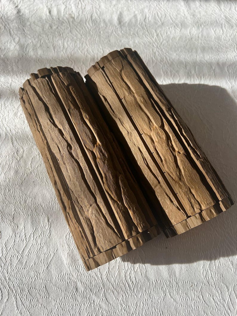May include: Two cylindrical wooden containers with a textured, bark-like appearance. The containers are a medium brown color and appear to be made of carved wood. The containers are lying on a white, textured surface.