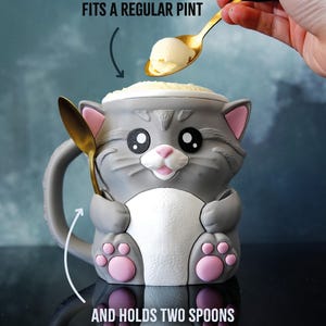 Op de afbeelding: Een grijze katvormige mok met roze accenten, ontworpen om twee lepels te bevatten. De mok heeft een witte buik en roze poten. Tekst luidt "FITS A REGULAR PINT" en "AND HOLDS TWO SPOONS". Een gouden lepel houdt een bolletje ijs.