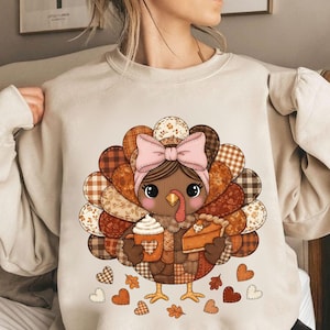 May include: Beige sweatshirt featuring a cartoon turkey with a pink bow, holding a pumpkin spice latte and a slice of pie. The turkey has a patchwork design with fall-themed patterns. Hearts and leaves are scattered around the turkey.